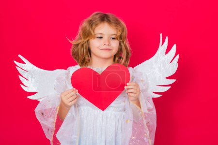 Foto de Angel kid with heart. Valentine angel. Little cupid child. Kid angel with angels wings, isolated on red studio background. Valentines day gift card - Imagen libre de derechos