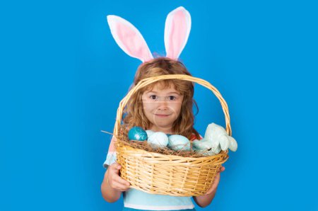 Foto de Conejito con cesta de Pascua aislado sobre fondo azul. Niño con orejas de conejo cazando huevos de Pascua. Caza de huevos de Pascua. Cara divertida de los niños - Imagen libre de derechos