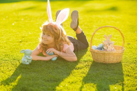 Téléchargez les photos : Child laying on grass in park wit easter eggs. Child with easter eggs and bunny ears laying on grass. Happy Easter kids face - en image libre de droit