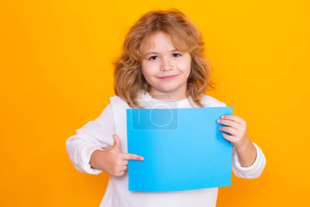 Foto de Kid boy with sheet of paper, isolated on yellow background. Portrait of a kid holding a blank placard, poster. Empty board for templates banner, copy spase, mock up - Imagen libre de derechos