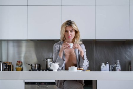 Photo for Beautiful woman drink coffee near table on kitchen at home. Sexy model covering big brest with pajama. Housewife on the kitchen in the morning - Royalty Free Image
