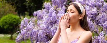 Foto de Spring allergy, flu. Banner. Sick woman sneezing covering nose with a wipe in a park. Spring allergy concept. Fashionable youth style. Allergic people. Among blooming trees and flowers in park - Imagen libre de derechos
