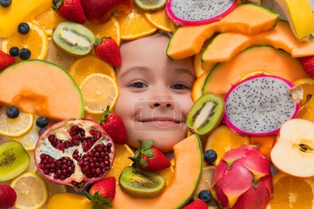 Photo for Happy smiling child face with fruits. Kids face with mix of strawberry, blueberry, strawberry, kiwi, dragon fruit, pomegranate, orange and melon. Assorted mix of fruits near child face - Royalty Free Image