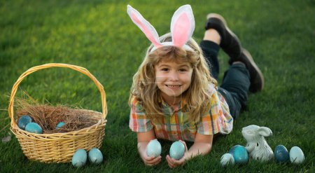 Foto de Bunny kids with rabbit bunny ears. Child hunting easter eggs in backyard laying on green grass. Panoramic web banner frame - Imagen libre de derechos