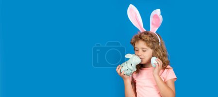 Téléchargez les photos : Bunny child. Child boy hunting easter eggs. Child boy with easter eggs and bunny ears isolated on blue background. Wide photo banner for website header design - en image libre de droit