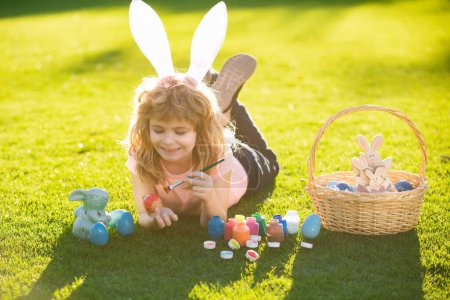 Téléchargez les photos : Child laying on grass in park wit easter eggs. Child boy in rabbit costume with bunny ears painting easter eggs on grass in spring park - en image libre de droit