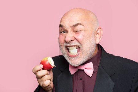 Téléchargez les photos : Mature man granddad in suit eat apple on pink background. Portrait of a funny elderly man eating an apple - en image libre de droit