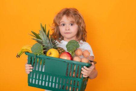 Téléchargez les photos : Food store. Child grocery cart, isolated studio yellow background with copy space. Little shopper. Kid holding shopping basket. Fresh organic, vegetables and fruits in grocery food store. - en image libre de droit