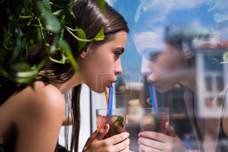 Foto de Beautiful woman hold cold beverage. Sensual woman drink cocktait with straw, looking out window. Close up portrait of sensual girl Female enjoys a summer drink - Imagen libre de derechos