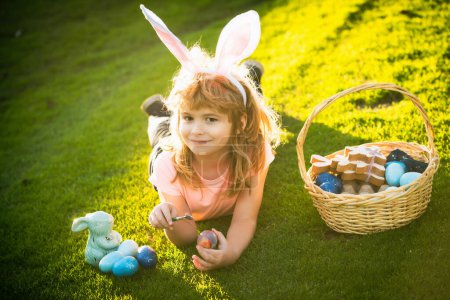 Foto de Niño con huevos de Pascua y orejas de conejo que ponen en la hierba. Chico gracioso, conejito de Pascua. Feliz cara de niños de Pascua - Imagen libre de derechos