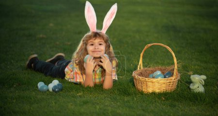 Téléchargez les photos : Child boy hunting easter eggs in spring lawn laying on grass. Bunny kids with rabbit bunny ears - en image libre de droit