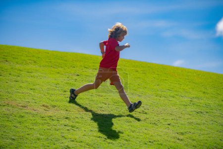 Foto de Sporty kid boy running on green grass and blue sky. Morning running with children. Child runner running in the nature. Morning jogging. Active healthy kids lifestyle - Imagen libre de derechos