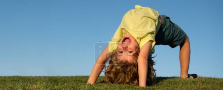 Téléchargez les photos : Joyeux petit enfant blond à l'envers allongé sur l'herbe dans le parc. Journée plus chaude pendant les vacances scolaires. Enfant incroyable garçon rêvant et souriant. Sport actif en plein air sain. Activité amusante - en image libre de droit
