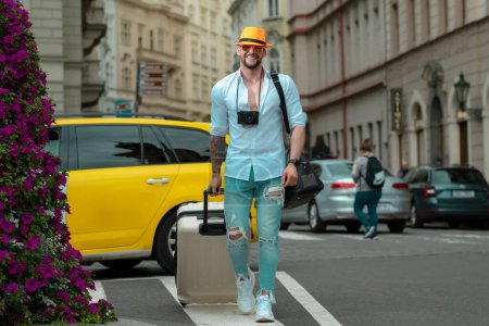 Foto de Traveller, fashion tourist. Male traveler with suitcase outdoor. Portrait of young caucasian man in hat and sunglasses on summer vacation - Imagen libre de derechos