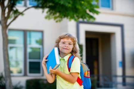 Foto de Un pequeño estudiante sonriente con mochila escolar y libro de ejercicios. Retrato de un alumno feliz fuera de la escuela primaria. Primeros planos del sonriente colegial hispano mirando a la cámara - Imagen libre de derechos