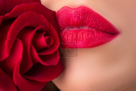 Téléchargez les photos : Beautiful young fashion woman with red lipstick. Glamour fashion model with bright gloss make-up posing at studio. CLoseup beauty portrait with a flower. Red rose. Beautiful woman lips with rose - en image libre de droit