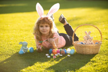 Foto de Kid laying on grass in park wit easter eggs. Child boy in rabbit costume with bunny ears painting easter eggs on grass in spring park - Imagen libre de derechos