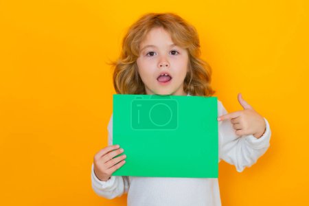 Photo for Funny kid showing index finger on green empty sheet of paper, isolated on yellow background. Portrait of a kid holding a blank placard, poster. Surprised face, amazed emotions of child - Royalty Free Image