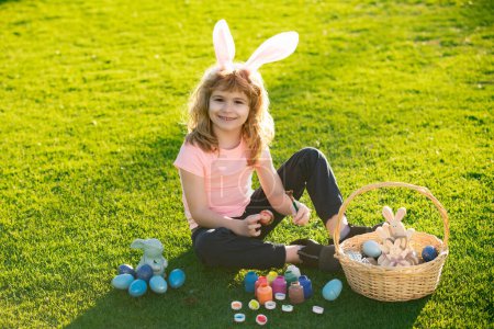Foto de Niño de Pascua en orejas de conejo cazando huevos de Pascua al aire libre. Lindo niño en traje de conejo con orejas de conejo que se divierten en el parque. Chico divertido, conejito de Pascua niños - Imagen libre de derechos