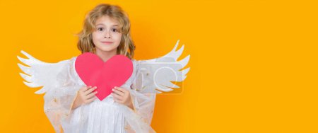 Foto de Angel child with heart. Valentines day. Blonde cute child with angel wings on a yellow studio background. Wide banner panoramic header. Happy angel child - Imagen libre de derechos