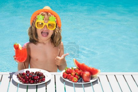 Foto de Excited kid with thumbs up on summer vacation. Kid with fruits and juice smoothie cocktail in summer pool. Child on summer vacation. Kid drink cocktail, strawberry smoothie in the pool - Imagen libre de derechos