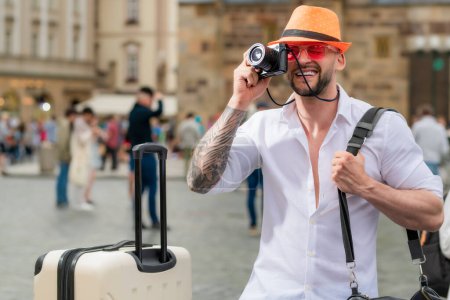 Foto de Tourist man in fashion hat jeans and sunglasses with suitcase walking on street. Man with travel suitcase on vacation. Man travel and adventure concept - Imagen libre de derechos