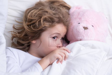 Foto de Child wakes up with toy teddy bear in the morning in the bedroom. Cute little boy waking up in bed - Imagen libre de derechos