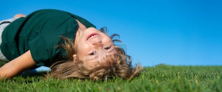 Foto de Child on spring nature background, horizontal photo banner for website header design. Sweet, happy child boy playing upside down on a grass in a park at a spring. Laughing, enjoying fresh. Funny kids - Imagen libre de derechos