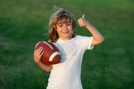 Téléchargez les photos : Child boy with american football, rugby ball. Kid boy playing with rugby ball in park. Cute portrait of a american football player, outdoor - en image libre de droit