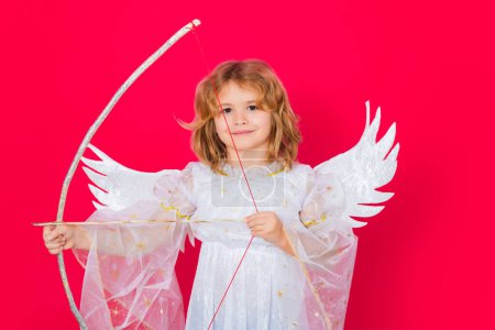 Téléchargez les photos : Angel kid shoots a love arrow from a bow on Valentines Day. Valentine angel. Little cupid child. Kid angel with angels wings, isolated on red studio background. Valentines day gift card - en image libre de droit
