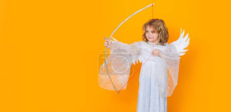 Téléchargez les photos : Angel kid shoots a love arrow from a bow on Valentines Day. Child at angel costume. Kid with angel wings. Isolated studio shot. Banner header with copy space - en image libre de droit