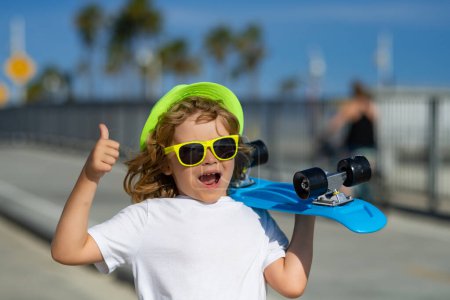 Foto de Happy boy laughing and holding skateboard outdoor. Excited child with thumb up, street portrait close up. Summer fashion kids portrait - Imagen libre de derechos