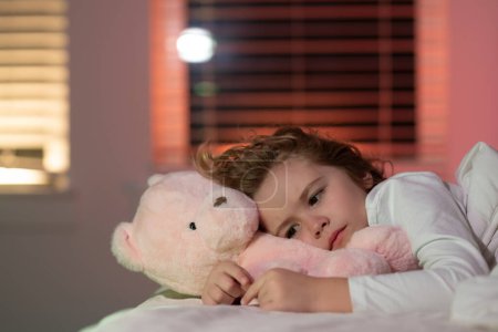 Photo for Cute boy trying to fall asleep with toy teddy bear. Child go to sleep in the night in the bedroom. Cute little boy asleep in bed - Royalty Free Image