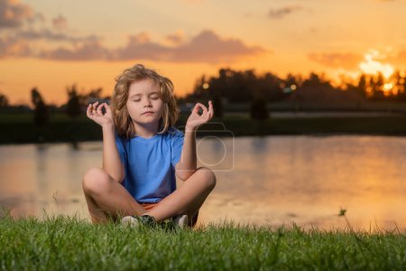 Téléchargez les photos : Child practices yoga and meditates in the lotus position near lake at sunset - en image libre de droit