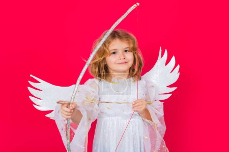 Téléchargez les photos : Angel kid shoots a love arrow from a bow on Valentines Day. Valentines day. Blonde cute child with angel wings on a red studio background. Happy angel child - en image libre de droit