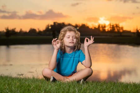 Foto de Kid meditating on lake at sunset. Yoga on nature. Mindfulness. Kid meditate in summer park. Calmness and relax, freedom concept. Harmony with nature. Freedom and carefree kids - Imagen libre de derechos