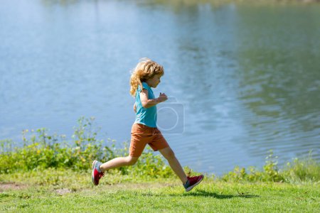 Foto de Kid boy running on green grass near lake in summer park. Sporty child boy runner running in summer park. Active kids, sport children - Imagen libre de derechos