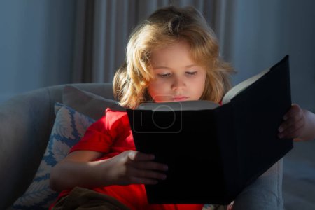 Téléchargez les photos : Kid boy reading book at living room. Close up portrait of cute blonde child reading interesting kids book story - en image libre de droit