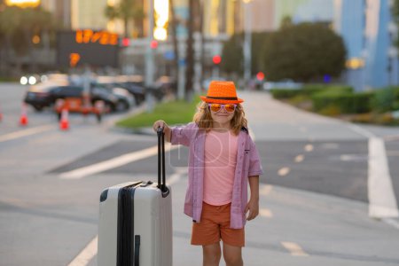 Foto de Funny child traveller. Child with suitcase going on vacation. Tourist boy on holiday, travel kids concept. Cute boy with suitcase on city street background - Imagen libre de derechos