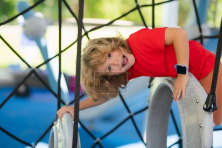 Foto de Child climbs up in a park on a playground on a summer day. Childrens playground in a public park. Recreation for children. Kid hanging at outdoor playground - Imagen libre de derechos