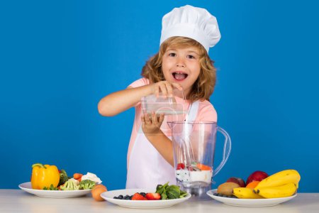 Téléchargez les photos : Child chef cook prepares milk shake smoothie in isolated blue studio background. Kids cooking. Teen boy with apron and chef hat preparing a healthy vegetables meal in the kitchen - en image libre de droit