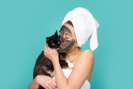 Téléchargez les photos : Woman with black cat apply charcoal facial mask mask isolated over blue studio background. Spa, cosmetic mask. Beautiful woman with black clay facial mud mask - en image libre de droit