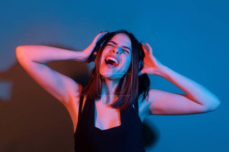 Foto de Chica DJ. Mujer joven emocionada escuchar música con auriculares, bailando. Chica escuchando música usando auriculares inalámbricos, bailando y cantando aislados sobre fondo azul neón - Imagen libre de derechos