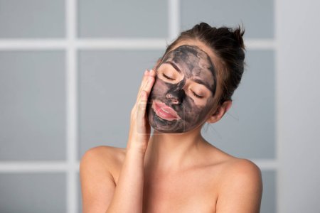 Téléchargez les photos : Femme appliquer masque facial charbon de bois isolé à la maison. Spa, masque cosmétique. Belle femme avec masque facial en argile noire. Soin de la peau, peeling, hydratation et soins de beauté - en image libre de droit
