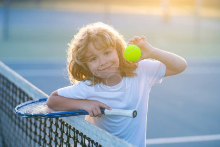 Téléchargez les photos : Enfant jouant au tennis sur le terrain extérieur. Le tennis est ma pièce préférée. Portrait d'un enfant assez sportif avec une raquette de tennis. Petit garçon souriant court de tennis. Entraînement et sport pour enfant - en image libre de droit