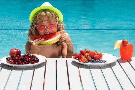 Téléchargez les photos : Un enfant mange de la pastèque. Enfant avec des fruits d'été dans la piscine. Enfant dans la piscine jouant et détendu dans l'eau. Vacances d'été avec les enfants - en image libre de droit