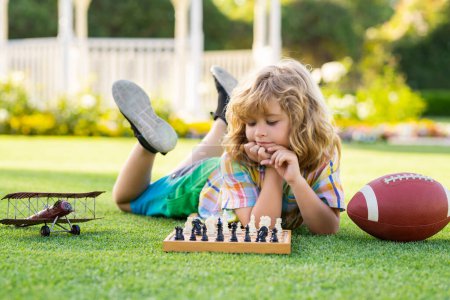 Téléchargez les photos : Concentrated child boy developing chess strategy, playing board game in backyard, laying on grass - en image libre de droit