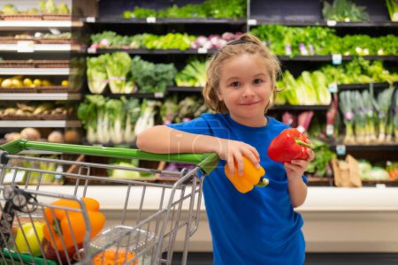 Foto de Kid with vegetables and peppers at grocery store. Kid at vegetable supermarket. Child choosing food in store or grocery store - Imagen libre de derechos