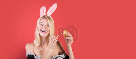 Foto de Happy young woman painting eggs on a red background. Cute bunny. Attractive young woman wearing bunny ears. Wide photo banner for website header design - Imagen libre de derechos