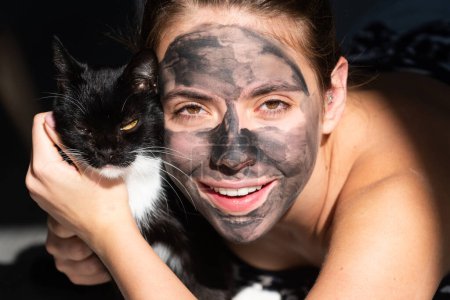 Téléchargez les photos : Morning with black cat. Young woman with naked shoulder and clay mask on face. Beautiful model applying cosmetic cream treatment on face. Facial treatment - en image libre de droit